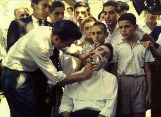 Έγρωμη φωτογραφία του 1954. Ο κουμπάρος ξυρίζει το γαμπρό παρουσία συγγενών και πολλών παιδιών. ΛΕΒΕΝΤΕΙΟ ΜΟΥΣΕΙΟ